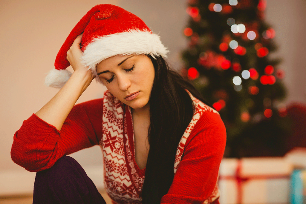 A woman with a Santa Claus hat on, has her eyes closed and is experiencing depression during the holidays.